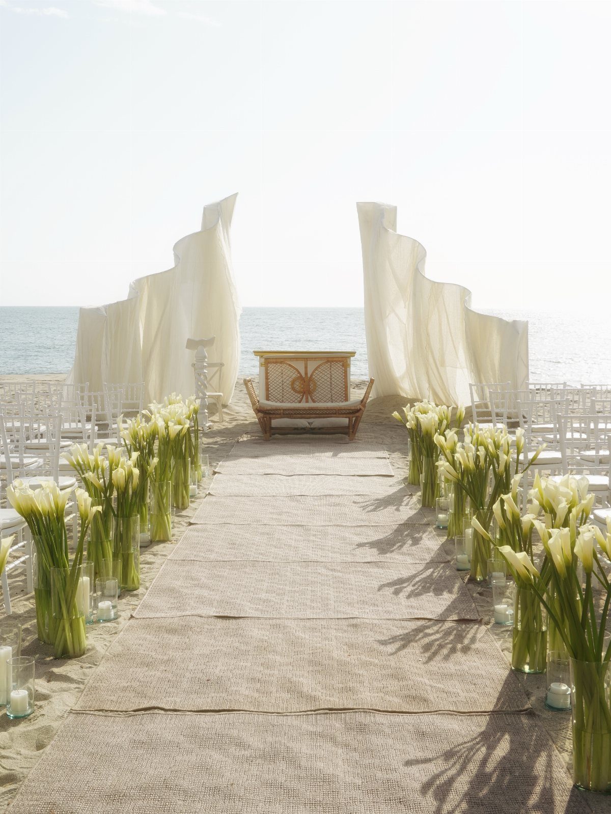 Beach wedding ceremony setup with burlap aisle, white tulips, and flowing white drapery.