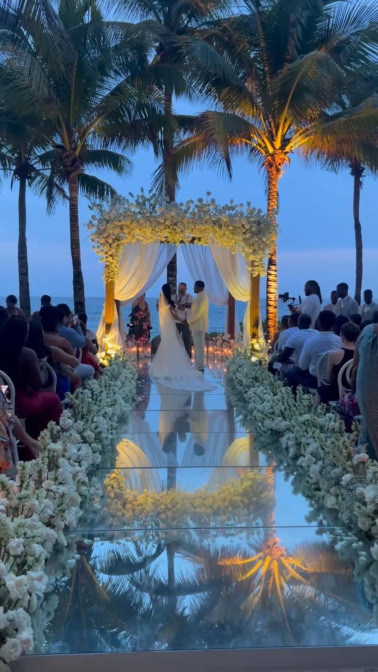 Wedding couple under a floral arch on a beach with a mirrored aisle.