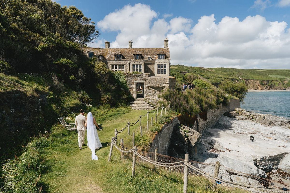 Bride and groom walking toward a historic stone house on a coastal cliffside.