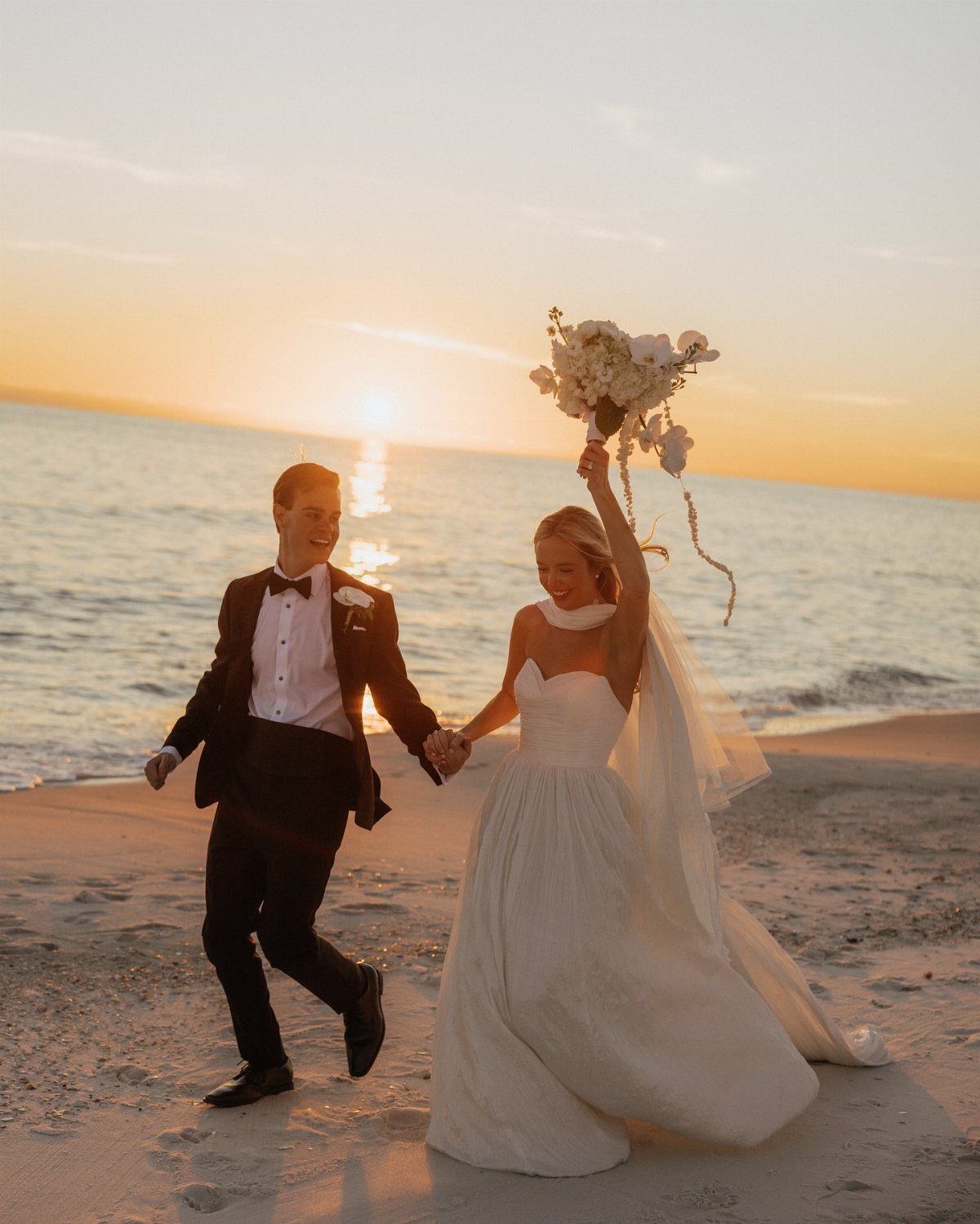 Bride and groom running on a beach at sunset during a wedding photoshoot.