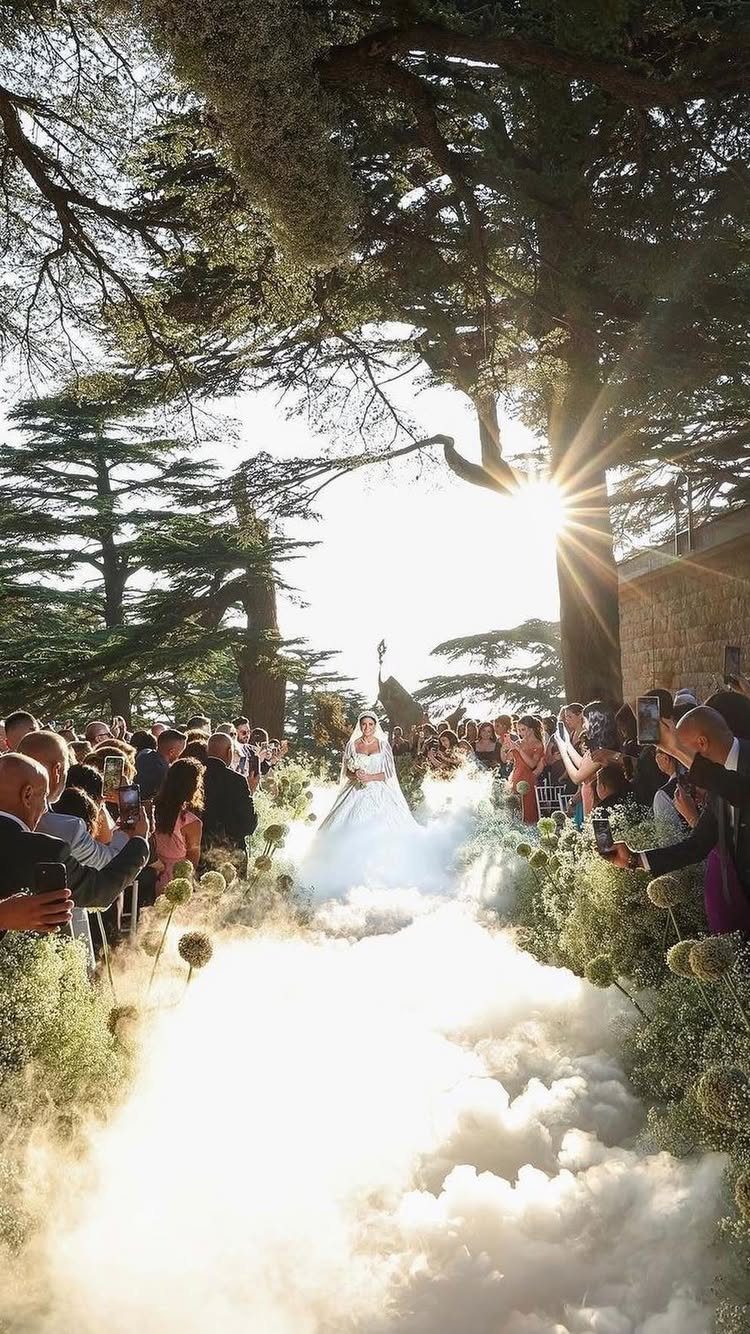 Bride walking down a wedding aisle filled with white fog under large trees