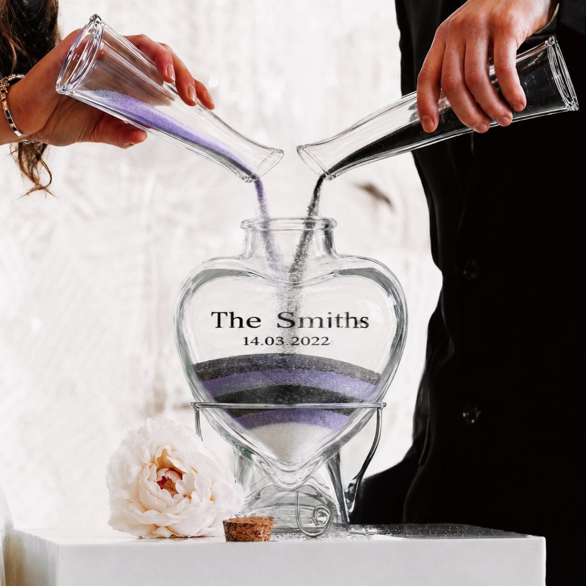 Two people pouring colored sand into a heart-shaped glass wedding jar.