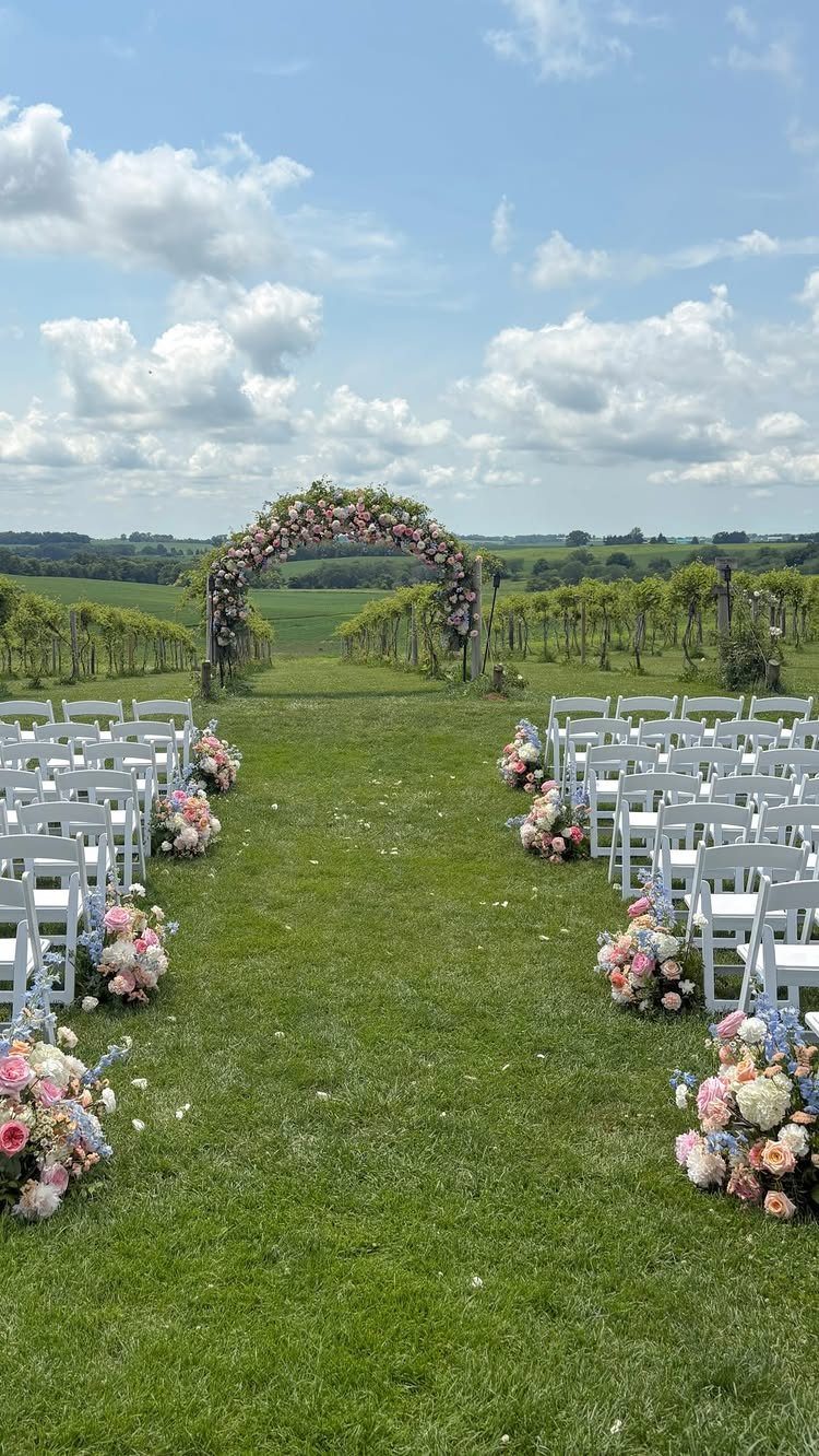 Outdoor wedding ceremony venue with white chairs, floral aisle markers, and a flower arch.
