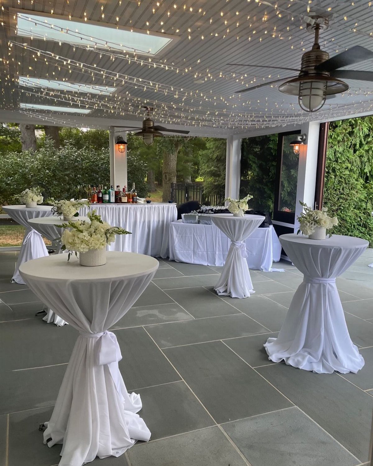 High-top cocktail tables with white linens and flowers under a lit patio ceiling.