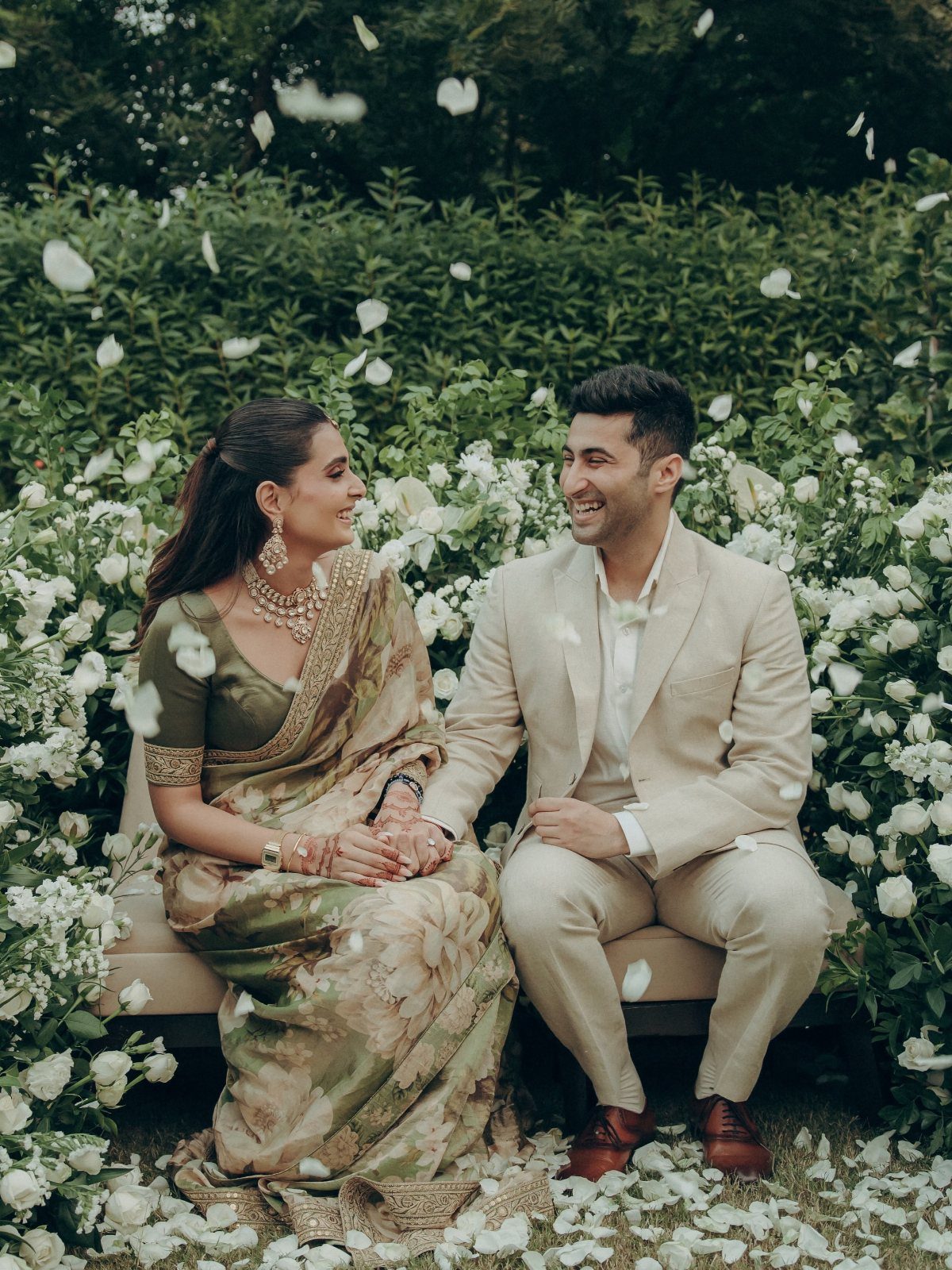 Couple in wedding attire sitting on a bench surrounded by white roses and petals