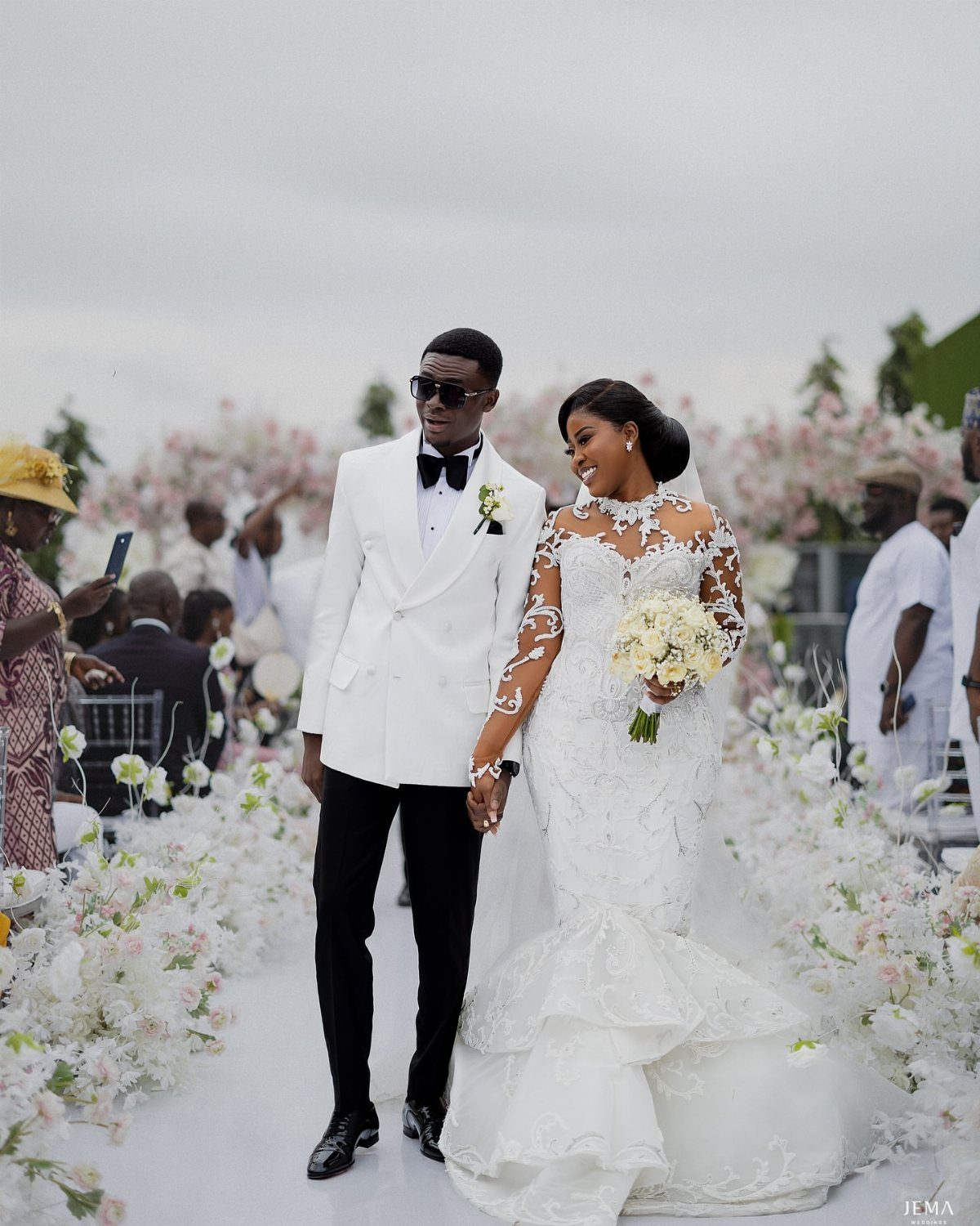 Bride in lace gown and groom in white tuxedo walking down flower-lined aisle.