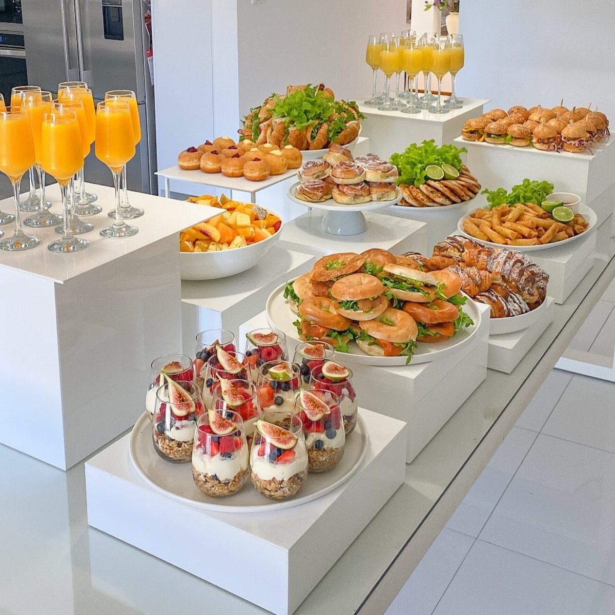 Breakfast spread with orange juice, fruit parfaits, and pastries on white pedestals.
