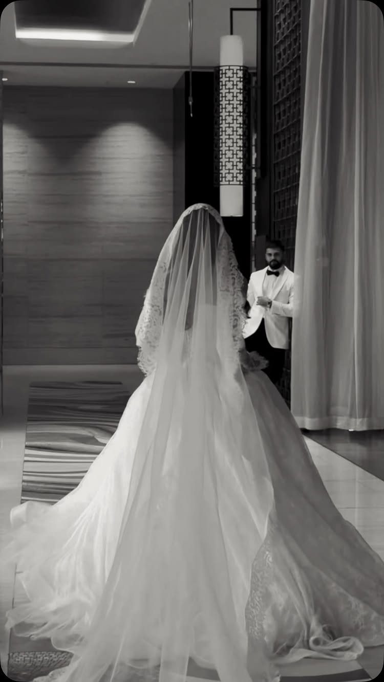 Bride in white gown and veil facing groom in white tuxedo jacket indoors.