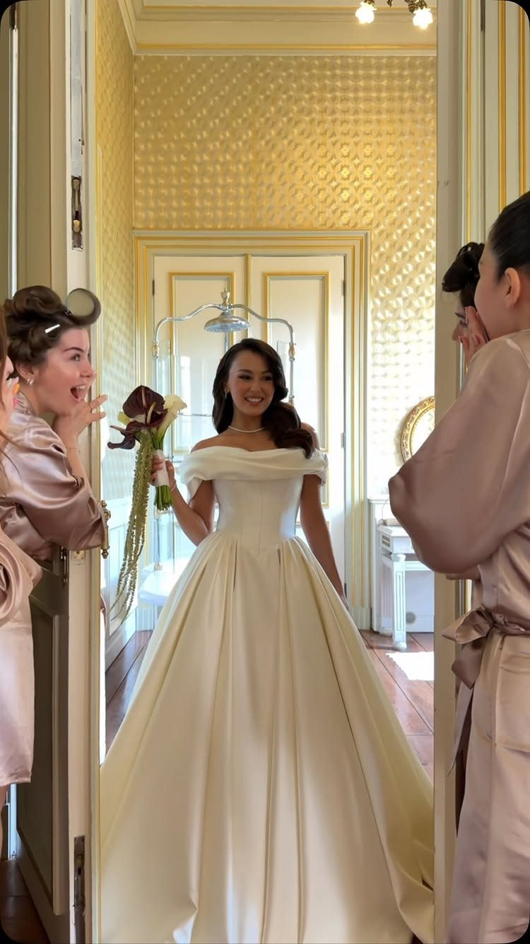Bride in a white ballgown holding flowers, viewed by bridesmaids in satin robes.