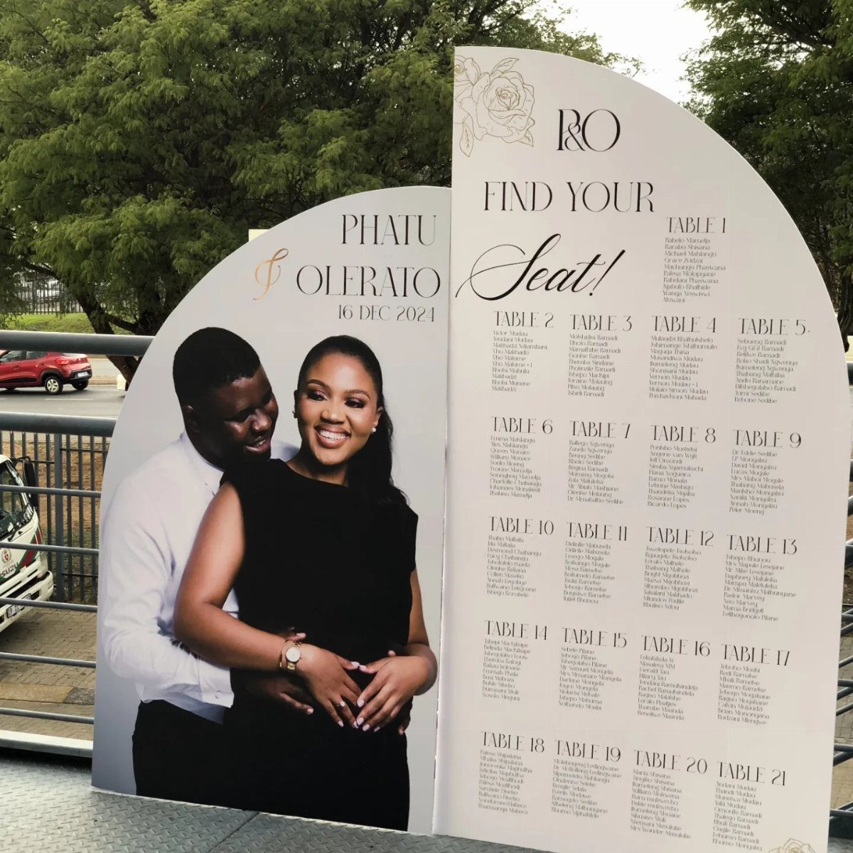 Curved white wedding seating chart with a couple's photo and guest table lists.