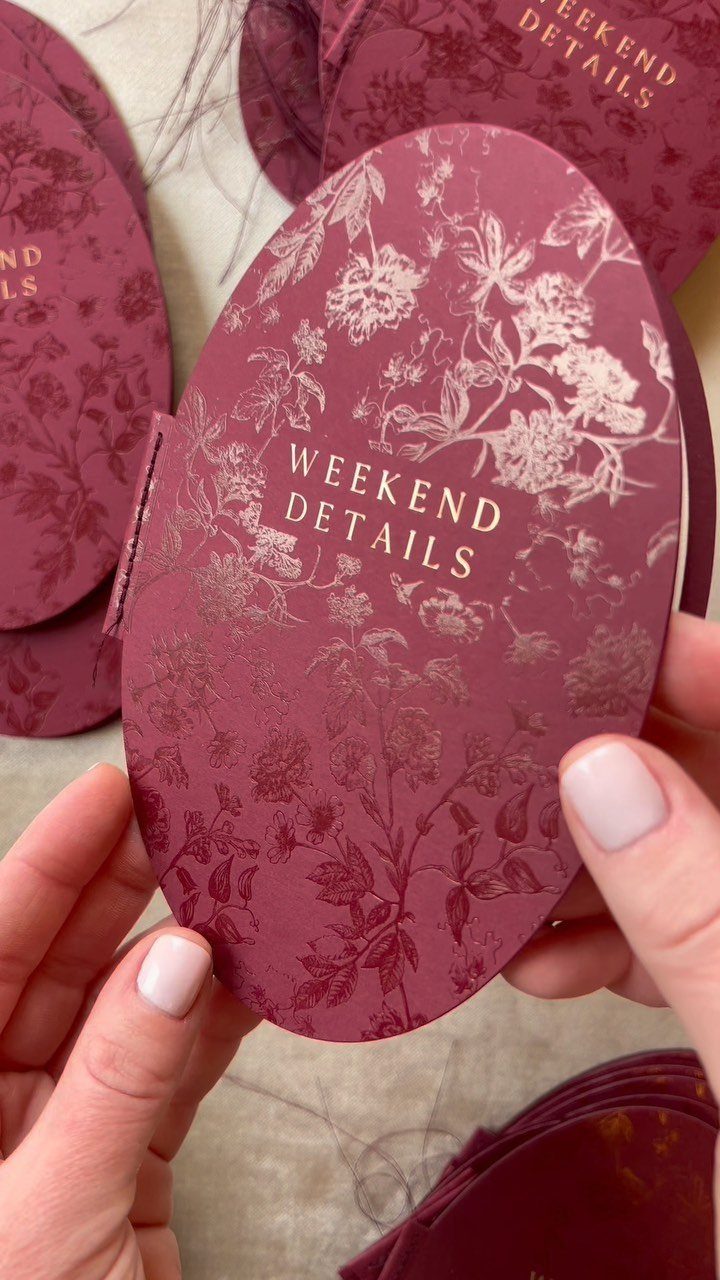 Burgundy oval wedding detail cards with gold floral patterns held by two hands.