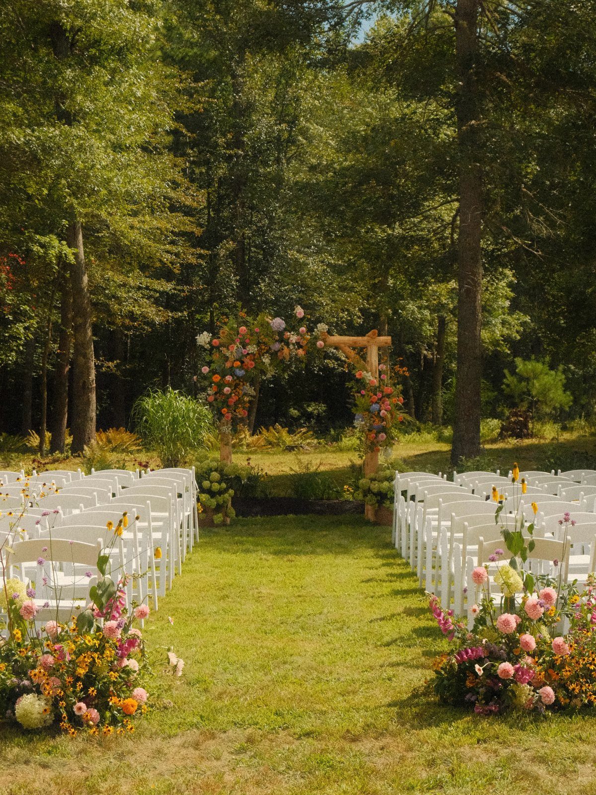 White wedding chairs and a floral arch on a lawn surrounded by trees