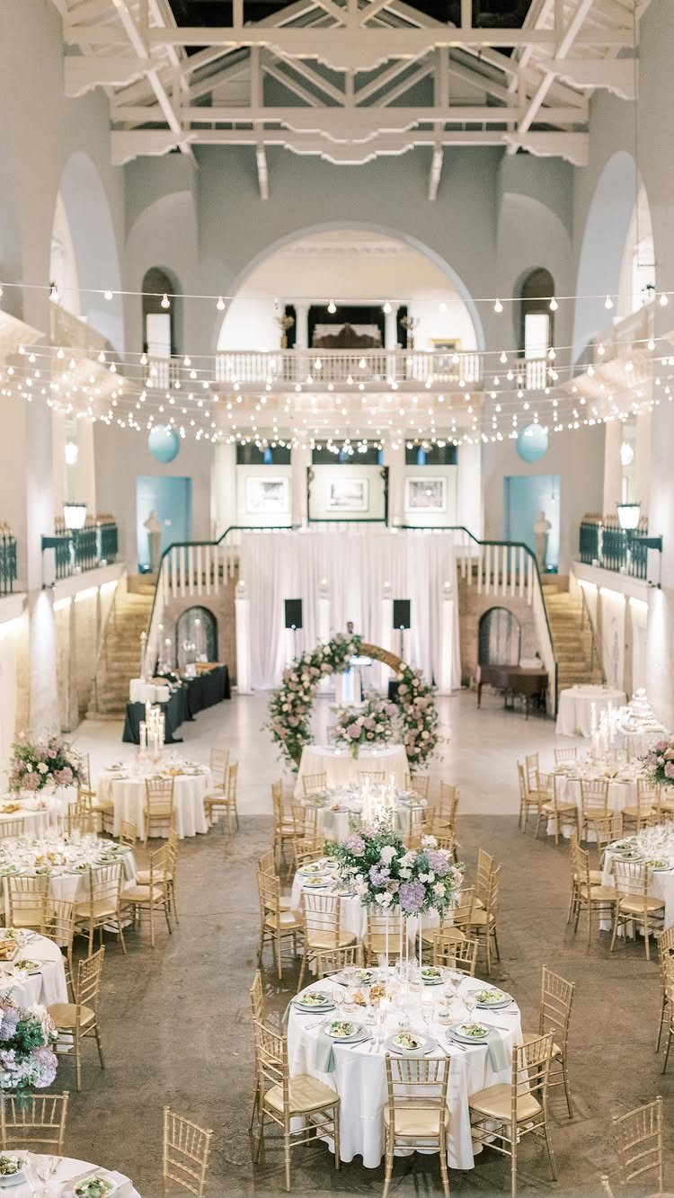 Wedding reception setup in a grand hall with white tables and floral arrangements.
