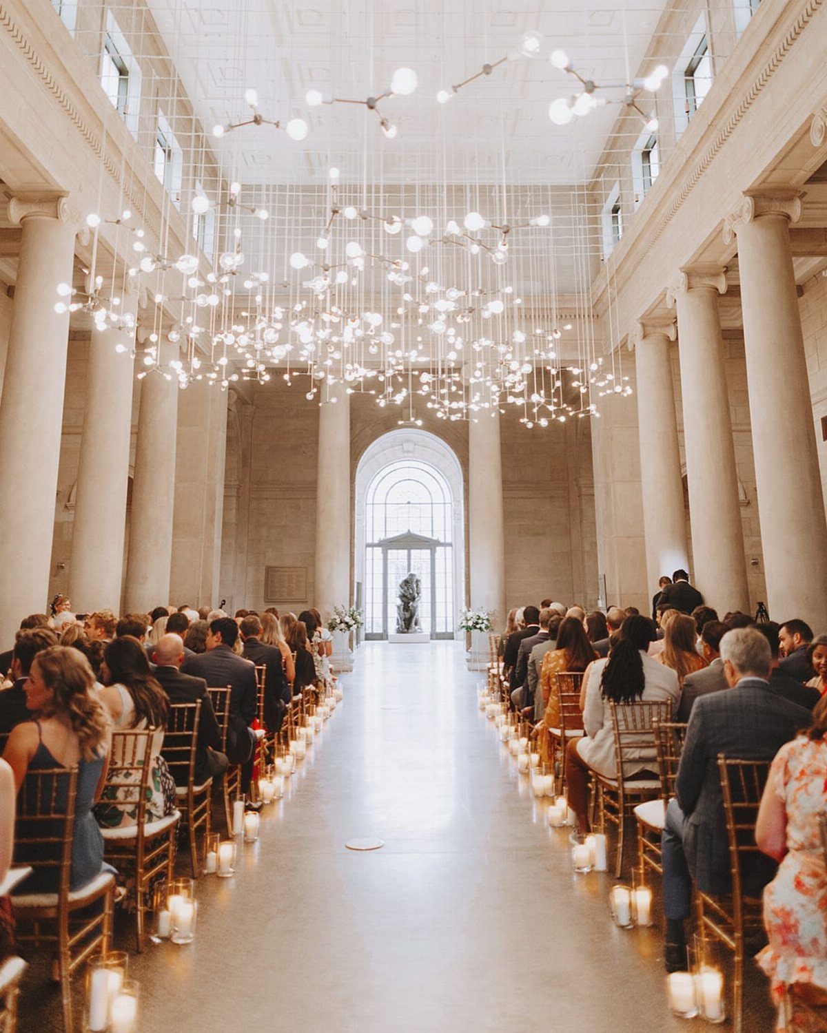 Wedding ceremony in a grand hall with stone columns and hanging globe lights.