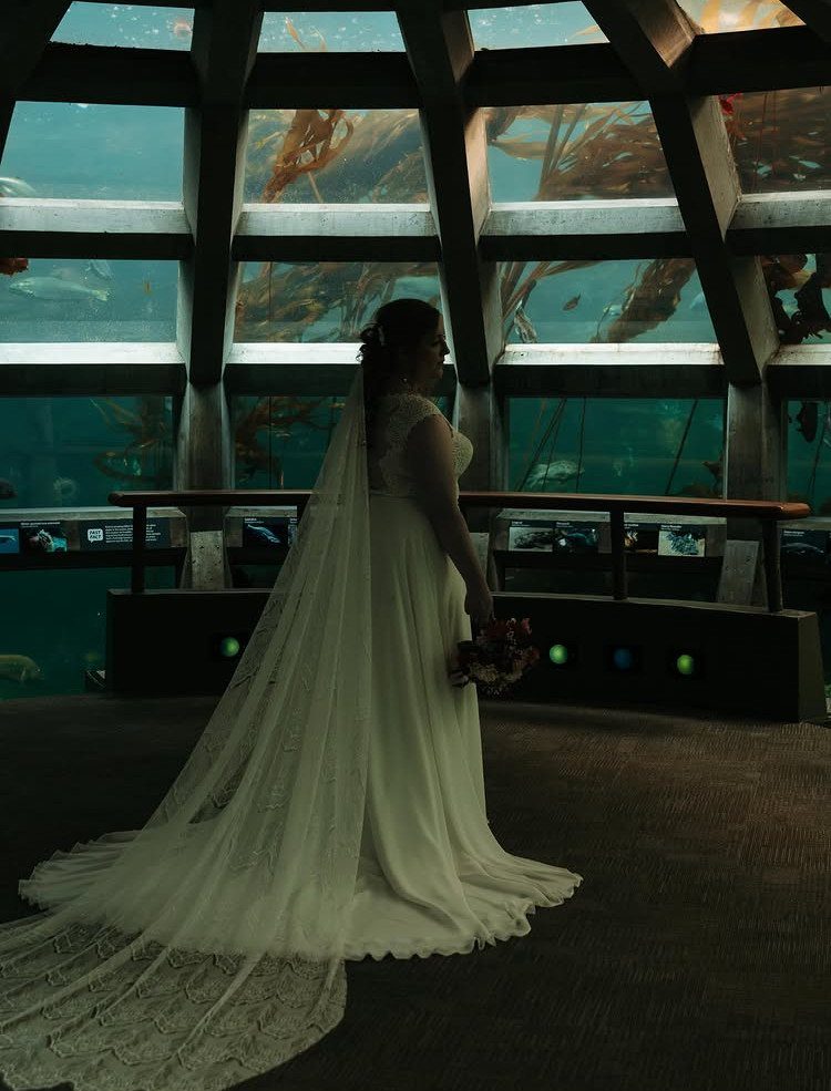 Bride in white gown standing before a large aquarium wall with marine life.