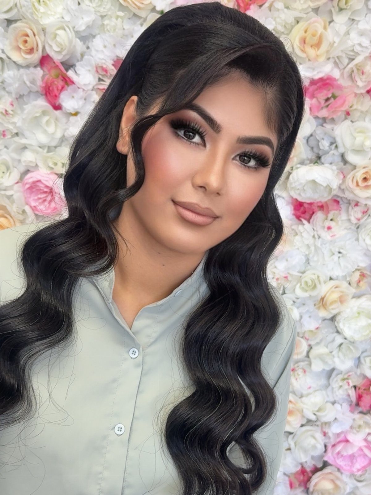 Woman with glamorous makeup and wavy black hair posing before a floral wall.