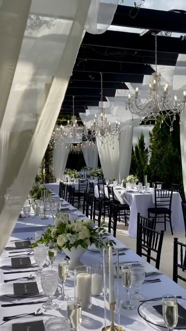 Outdoor wedding reception with long white tables, black chairs, and hanging crystal chandeliers.
