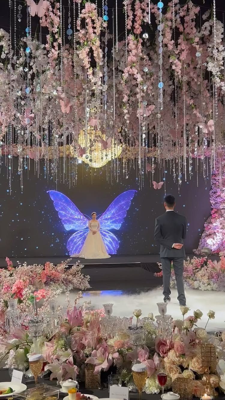 Groom facing bride on a stage with blue butterfly projection and floral decor