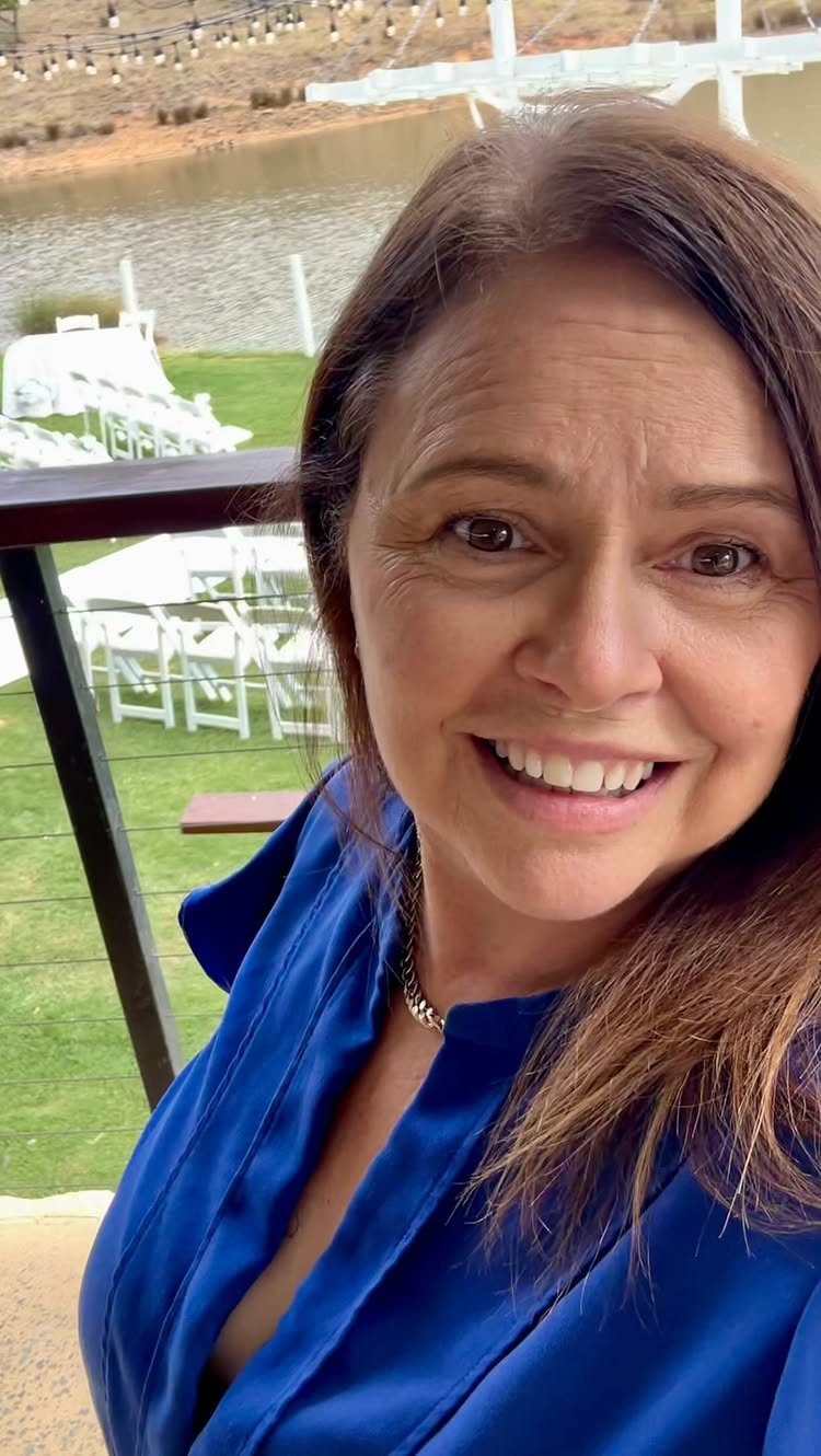 Smiling woman in a royal blue satin blouse overlooking an outdoor wedding setup.