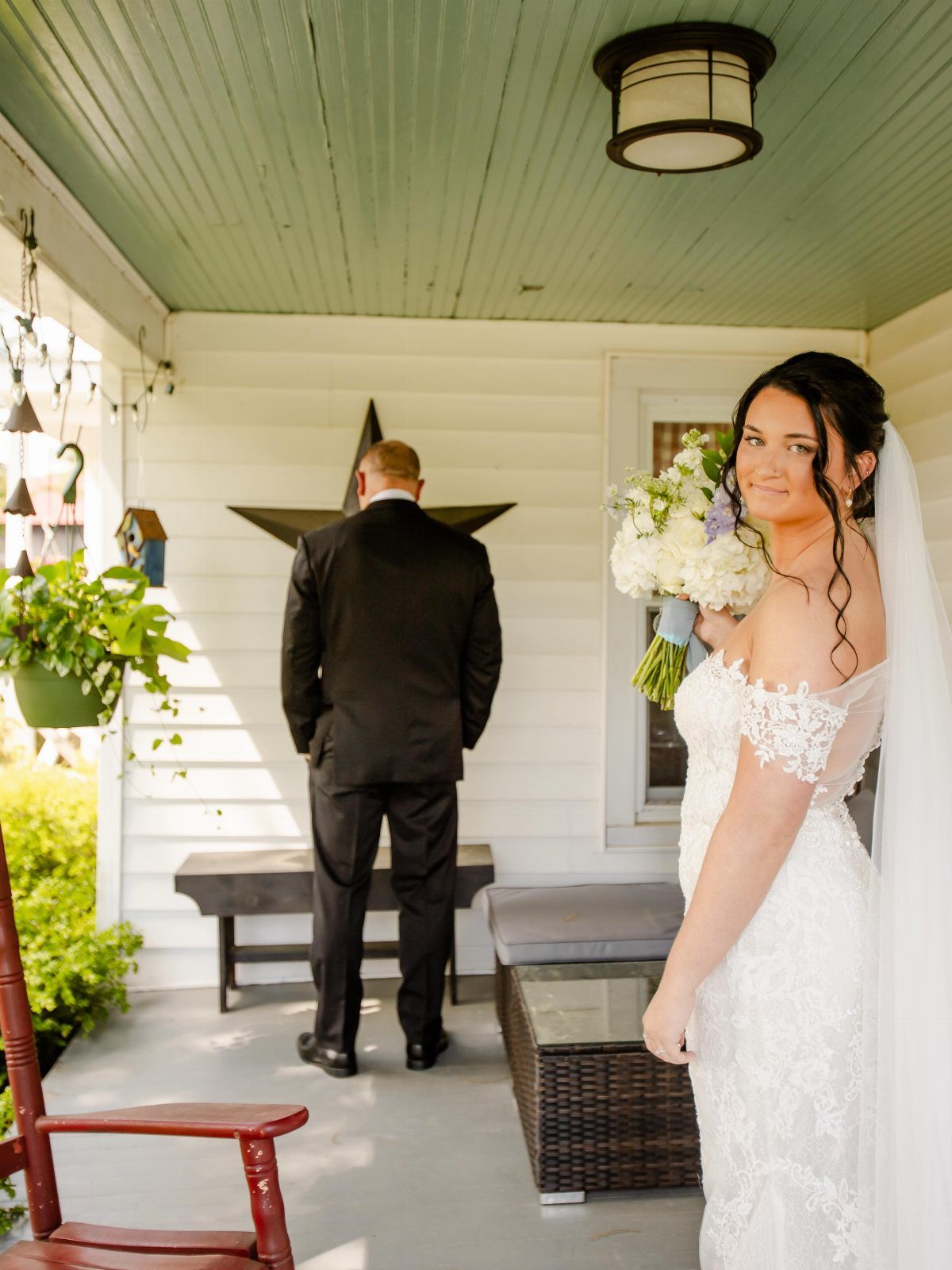 Bride in white lace dress standing behind a groom who has his back turned.