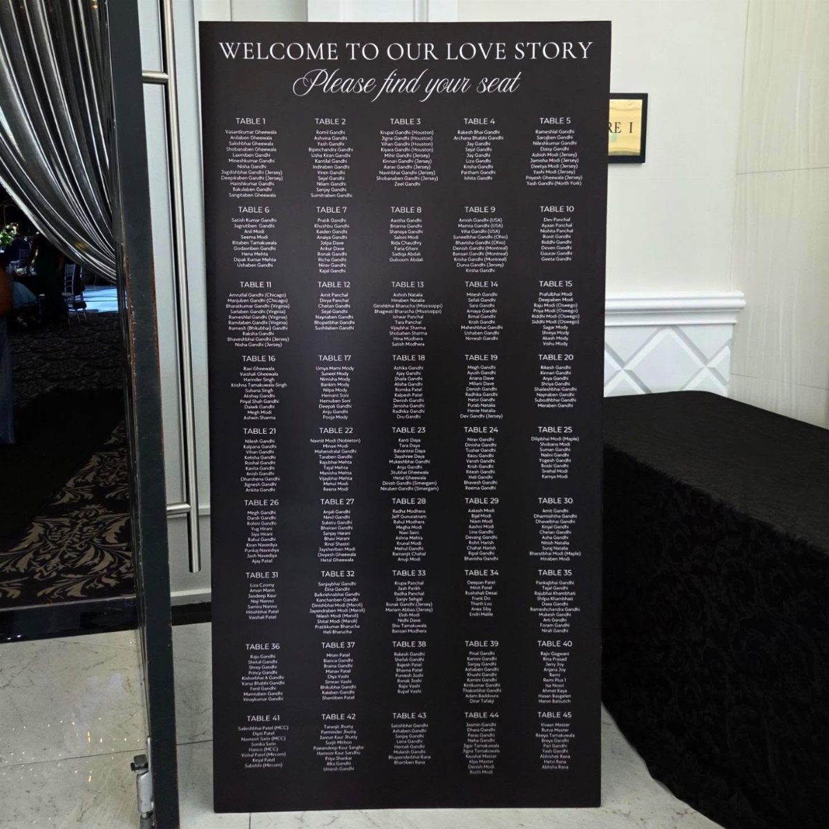 Black wedding seating chart board listing guests by table number in a white room.