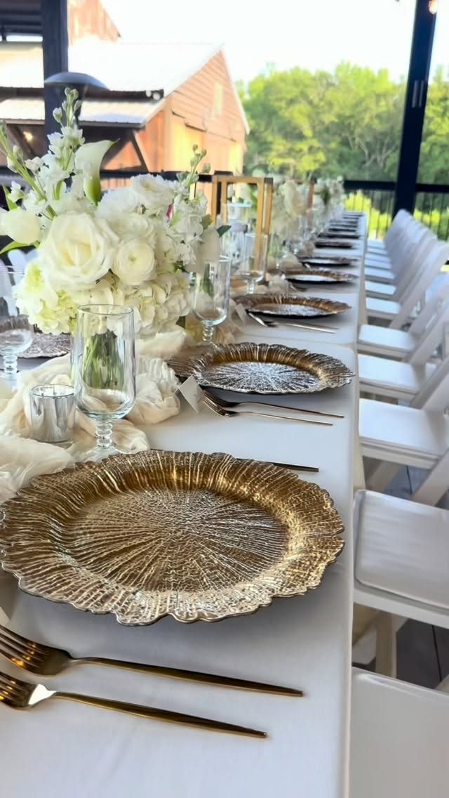Long wedding table set with gold charger plates and white floral centerpieces outdoors.