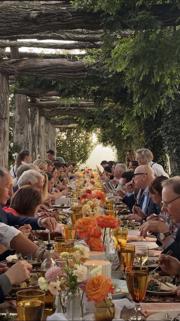 Guests dining at a long table under a vine-covered wooden pergola with floral centerpieces