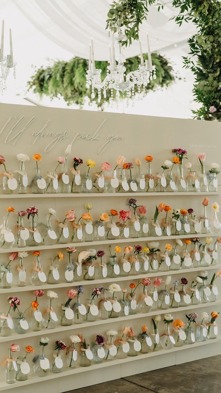 Seating chart with small glass bottles holding colorful flowers and name tags on shelves.