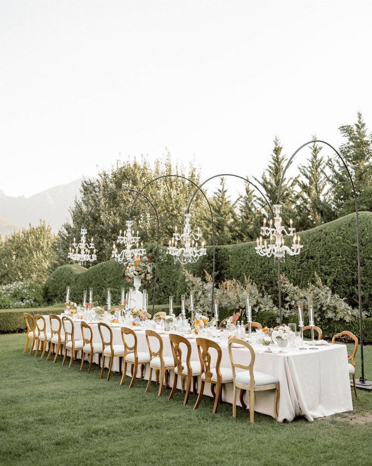 Long outdoor wedding banquet table with white linens, gold chairs, and hanging chandeliers.