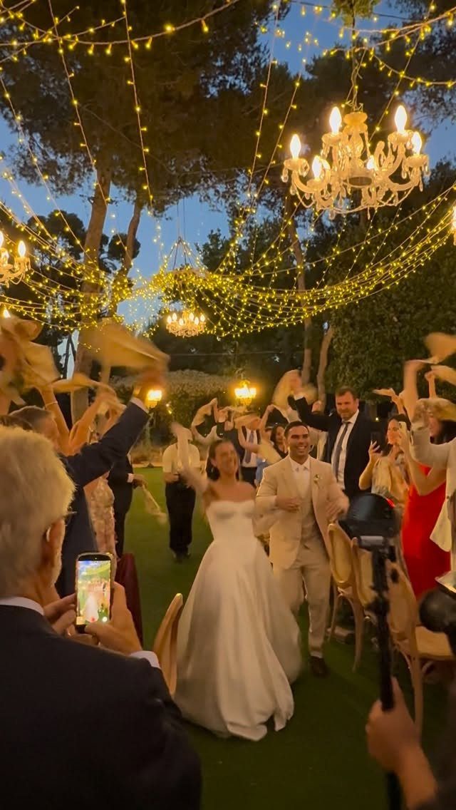Newlyweds walking through cheering guests under fairy lights at an outdoor wedding reception.