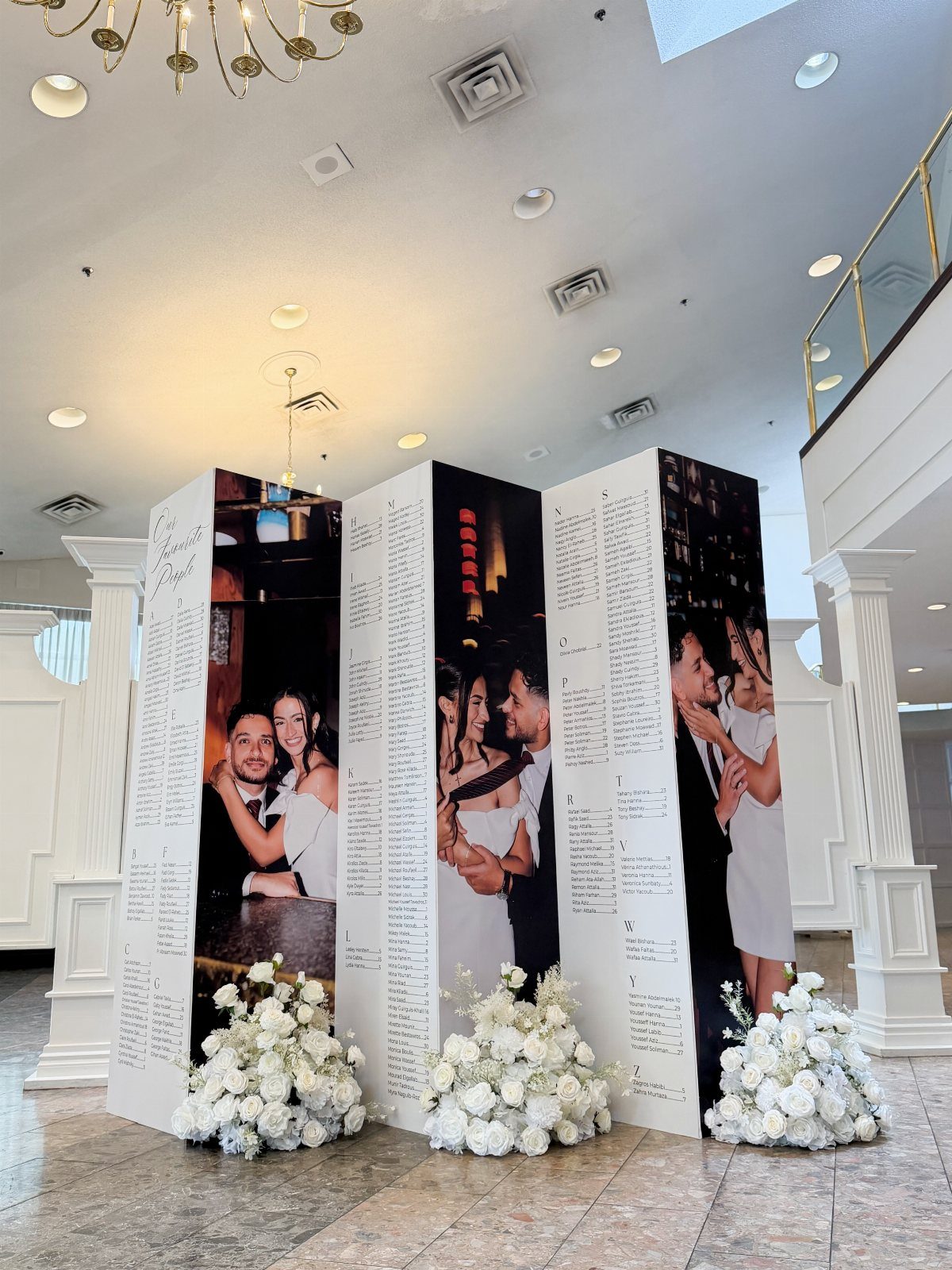 Three-panel wedding seating chart with couple photos and white floral arrangements at base.