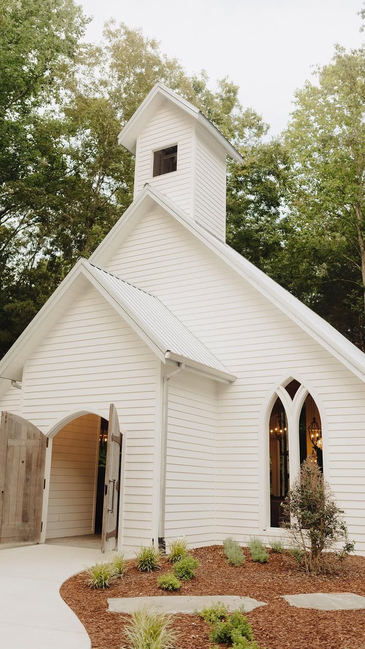 White wooden chapel with a steeple and arched windows surrounded by lush greenery
