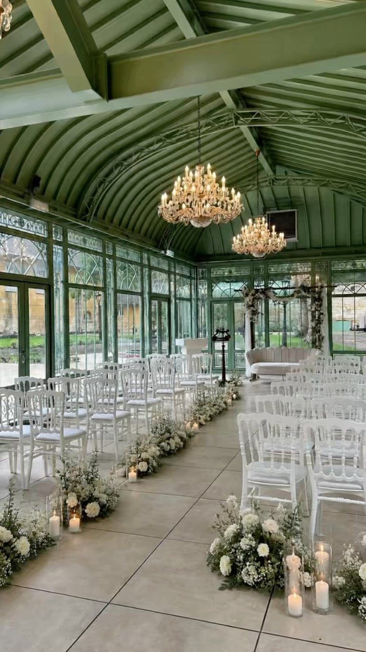 Wedding ceremony setup in a green glass conservatory with white chairs and floral aisle.