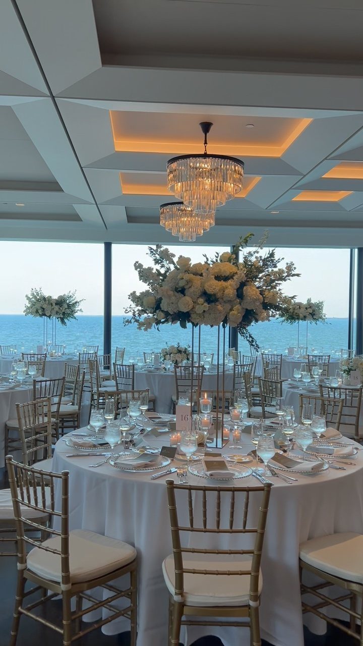 Wedding reception tables with white flowers and gold chairs overlooking a blue ocean.