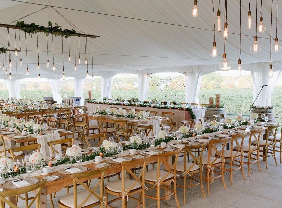 Wedding reception setup inside a white tent overlooking a lush green vineyard landscape.