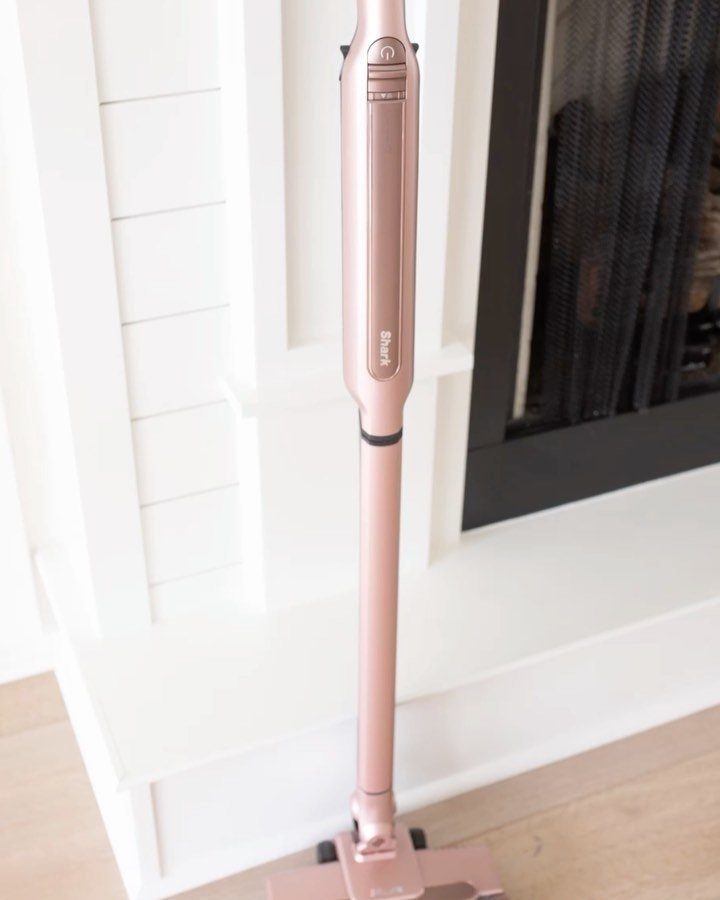 Rose gold cordless stick vacuum cleaner standing against a white paneled wall.