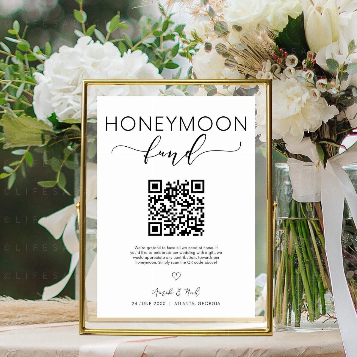 Gold-framed honeymoon fund sign with QR code surrounded by white floral arrangements.