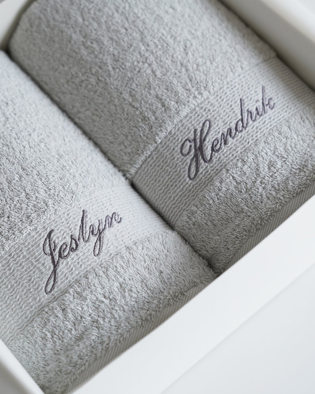 Two grey plush towels with embroidered names inside a white gift box.