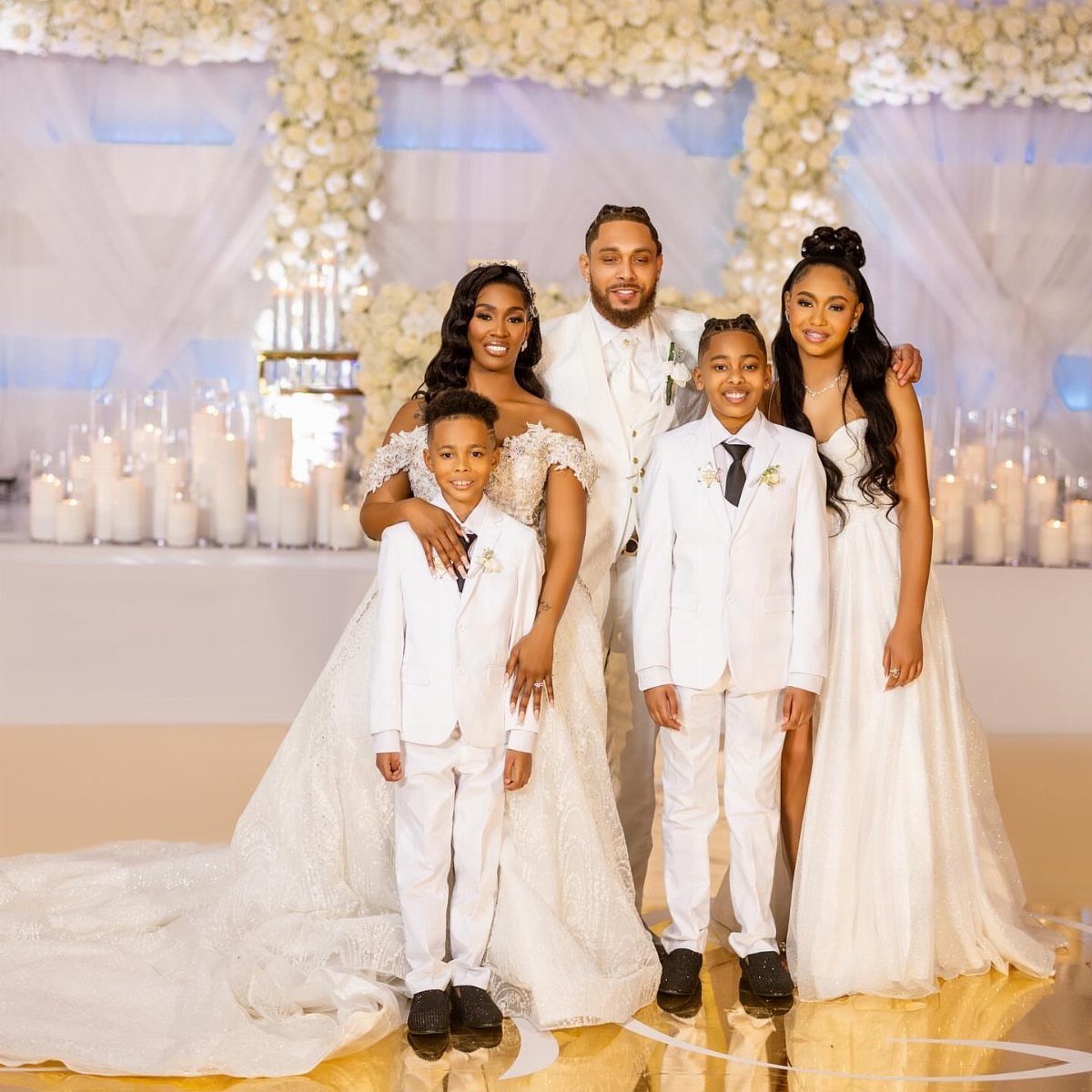 Family in formal white wedding attire posing before a floral and candle backdrop.