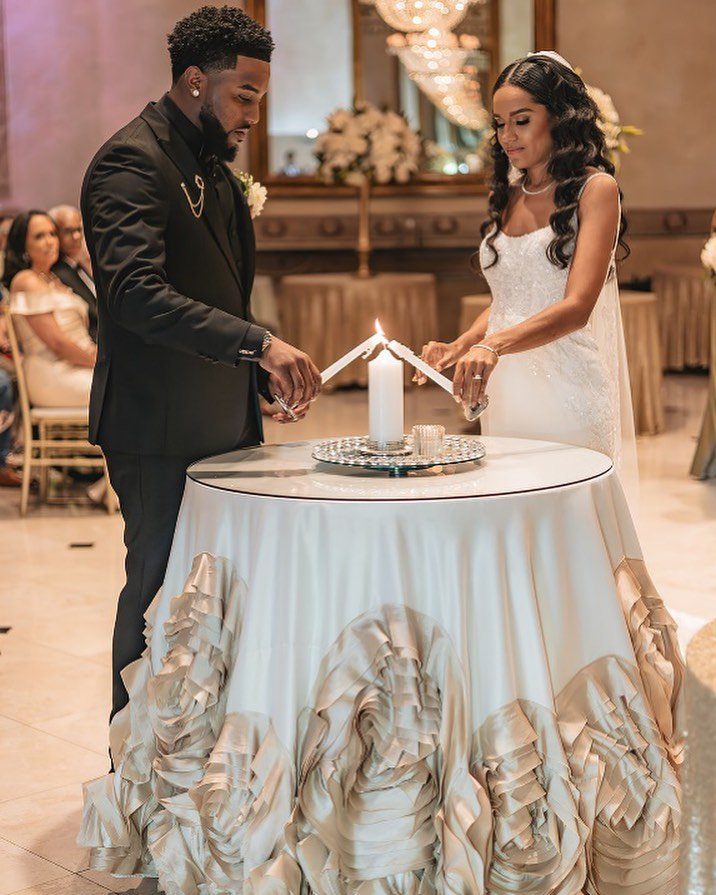 Couple in wedding attire lighting a unity candle at a decorated table.