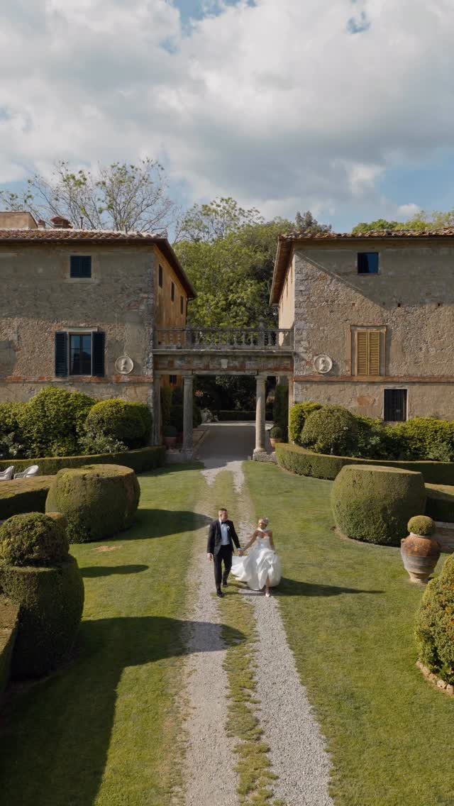Bride and groom walking down a gravel path between rustic stone Tuscan buildings.