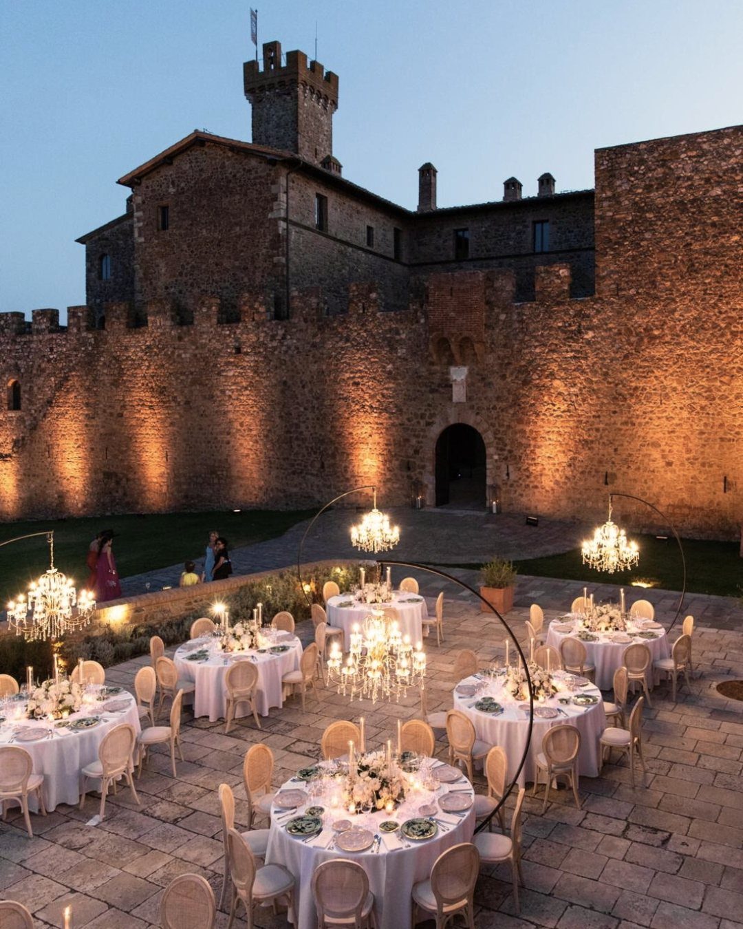 Wedding reception tables with chandeliers in a courtyard before a lit stone castle.