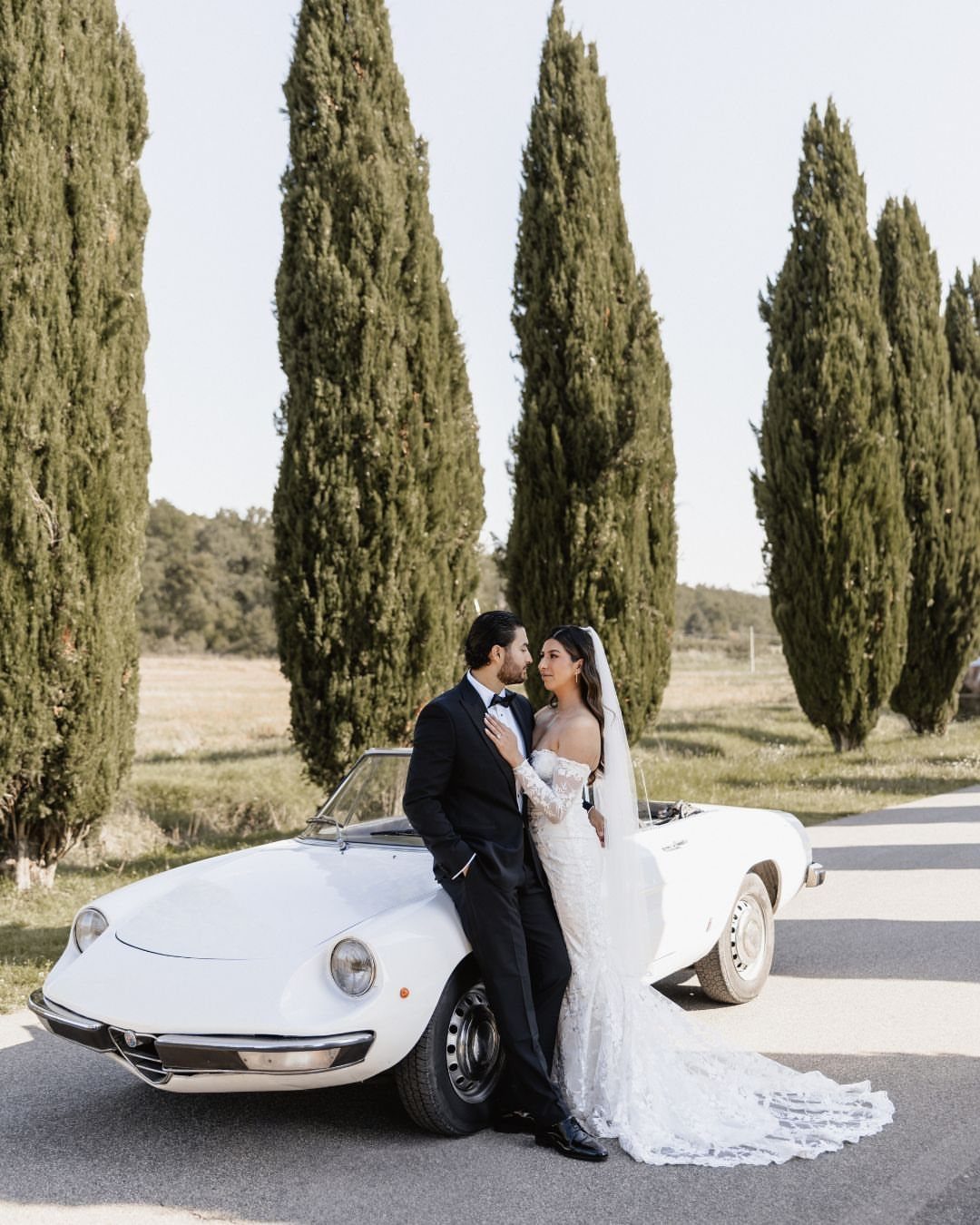 Bride and groom posing by a white vintage car among Tuscan cypress trees