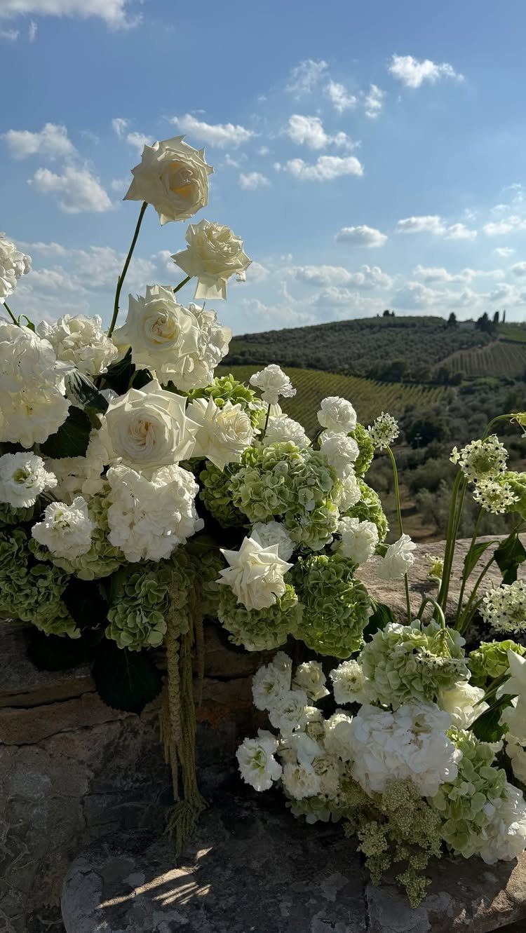 White roses and hydrangeas arrangement overlooking rolling green Tuscan hills under blue sky