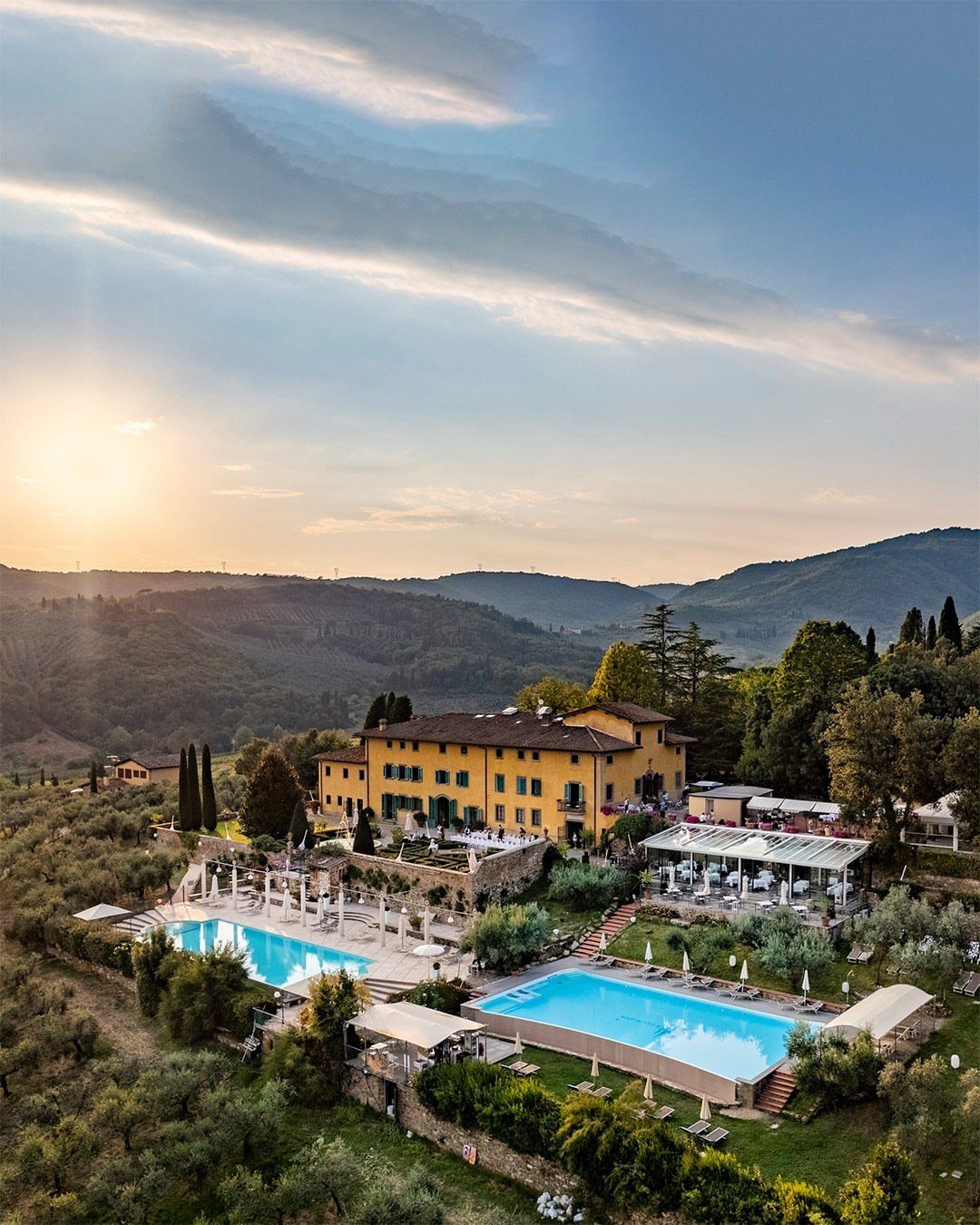 Yellow historic villa with two blue swimming pools overlooking rolling Tuscan hills.