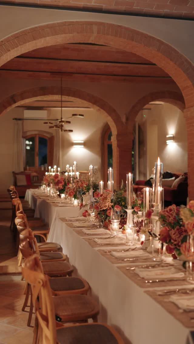 Long wedding table with white linens, pink flowers, and lit candles under brick arches.