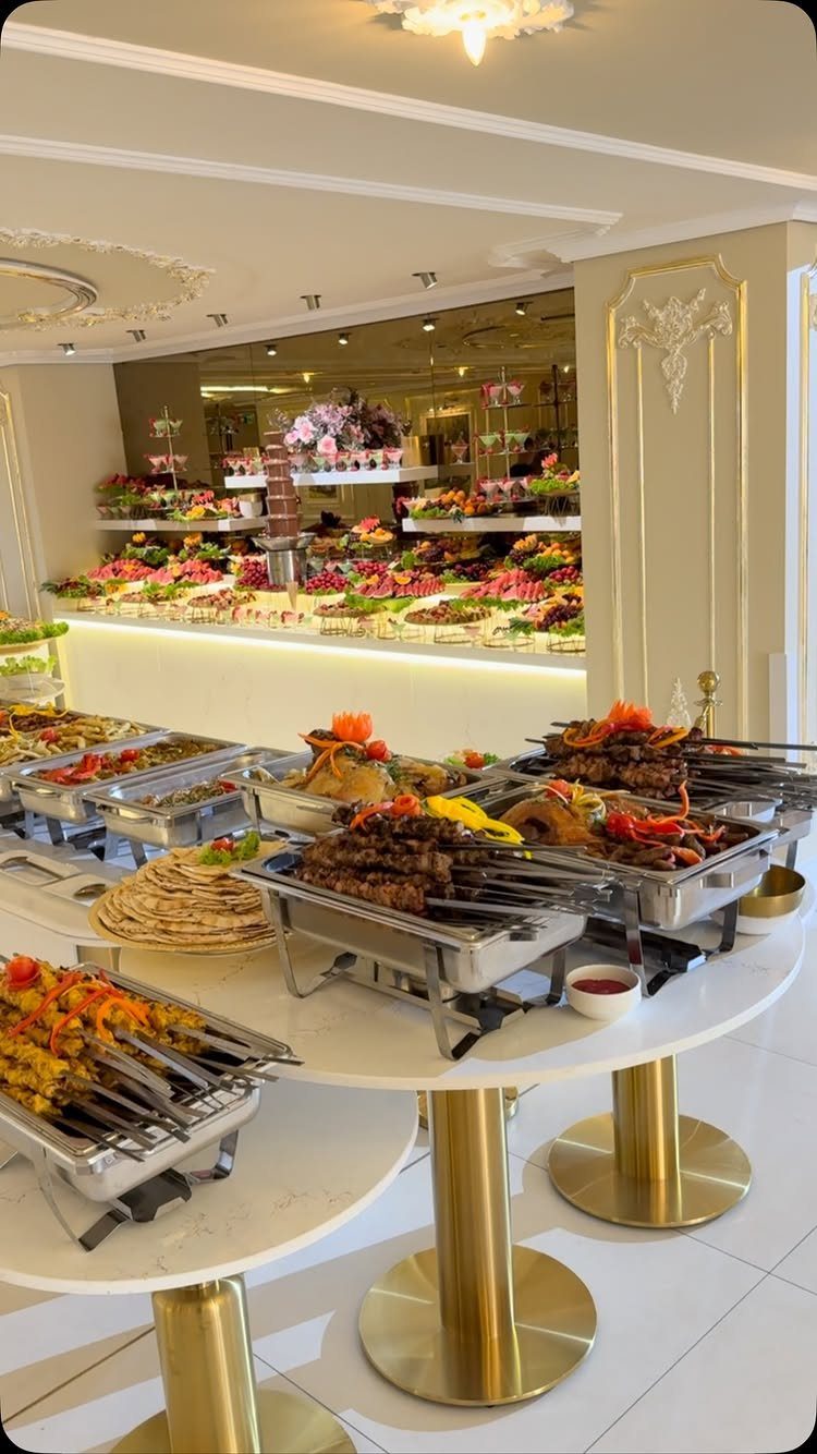 Buffet spread with meats and vegetables in silver chafing dishes on marble tables.