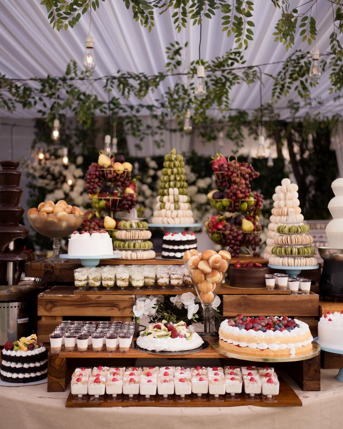 Elaborate dessert table with fruit towers, cakes, and macarons under a white tent.