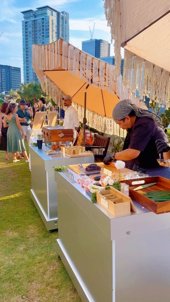 Chef preparing food at silver outdoor food stations under beige umbrellas with city skyline.
