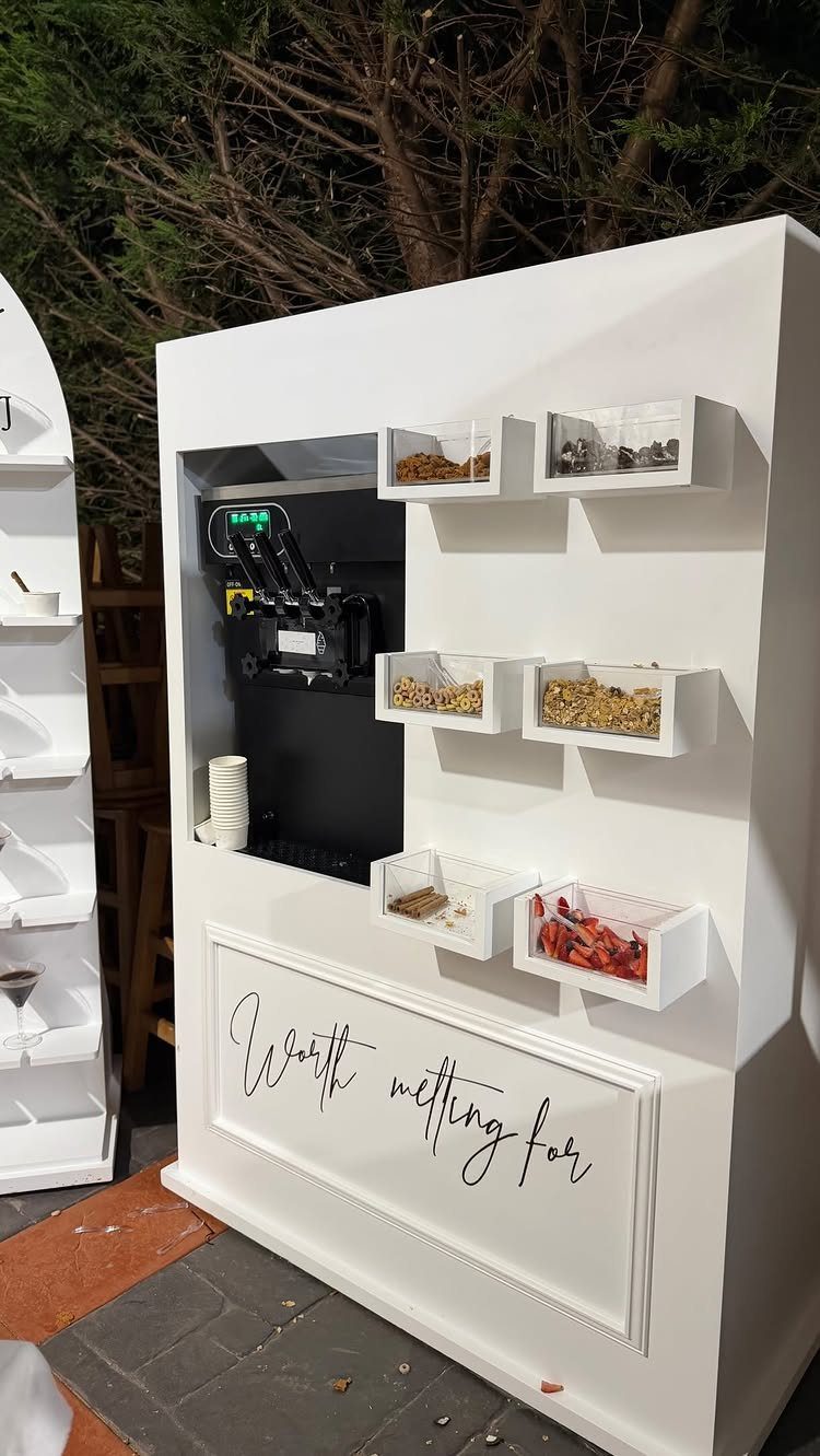 White dessert station with soft-serve machine and topping bins for guest customization.